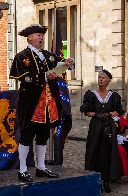 A town crier, wearing Georgian era regalia, spreads the news from a stand in the town square. 